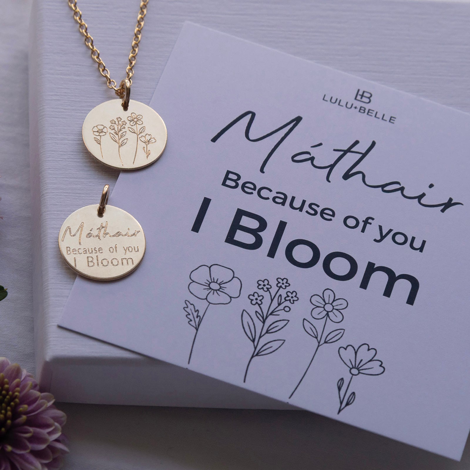 The Mathair Bloom Necklace shown front and back on a gift box, with a meaning card explaining the “Because of you I bloom” engraving.