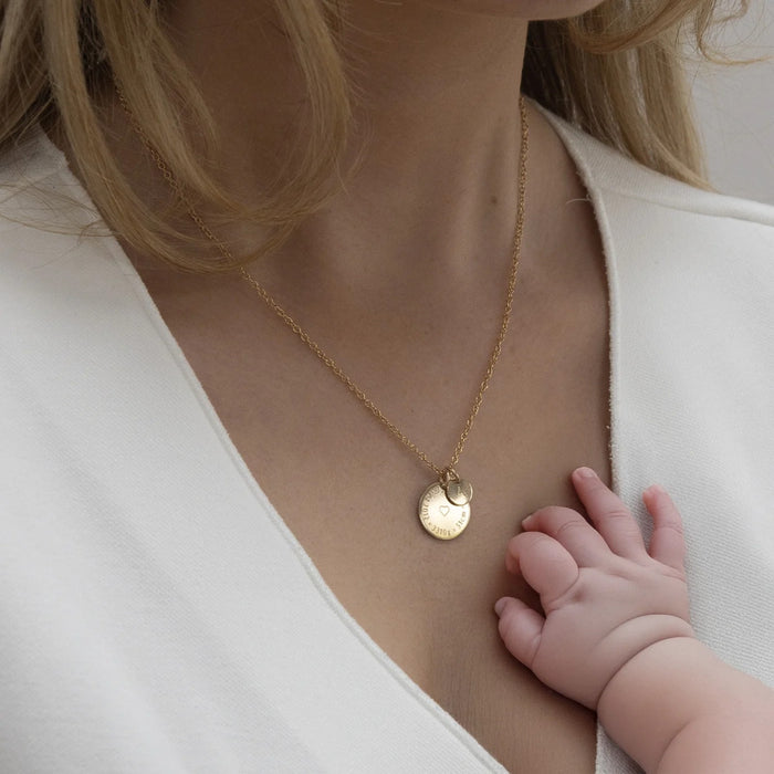 Gold Birthcoin necklace engraved with birth details, worn by a mum with her baby's hand on her chest